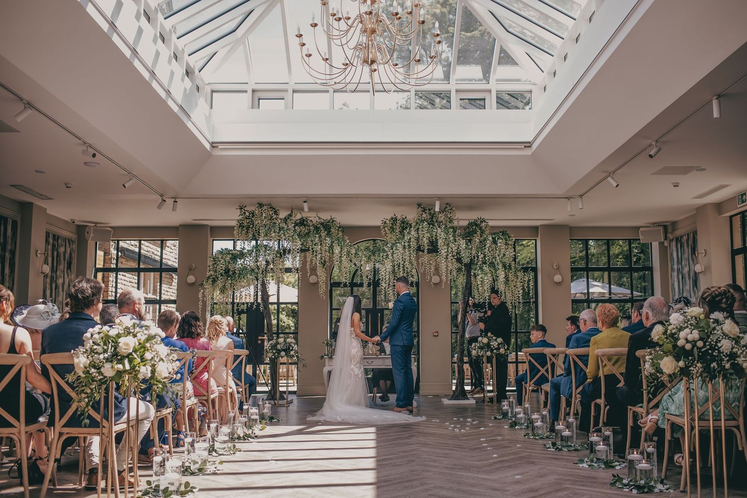 Bright wedding ceremony under glass skylight roof with floral arch and seated guests in aisle.