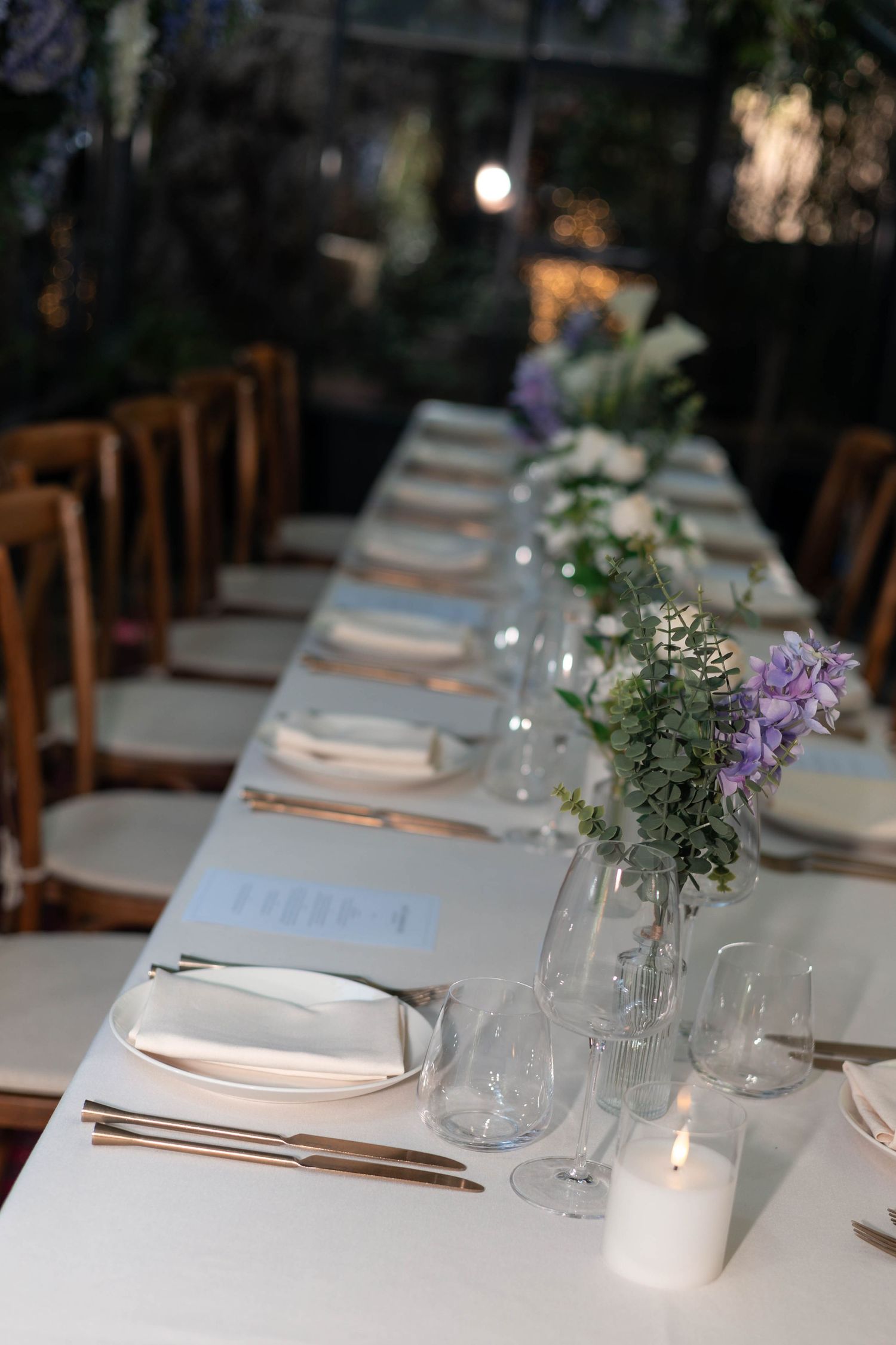 Elegant long dining table set with white linens, crystal glasses, candles, and purple floral arrangements for a formal event.