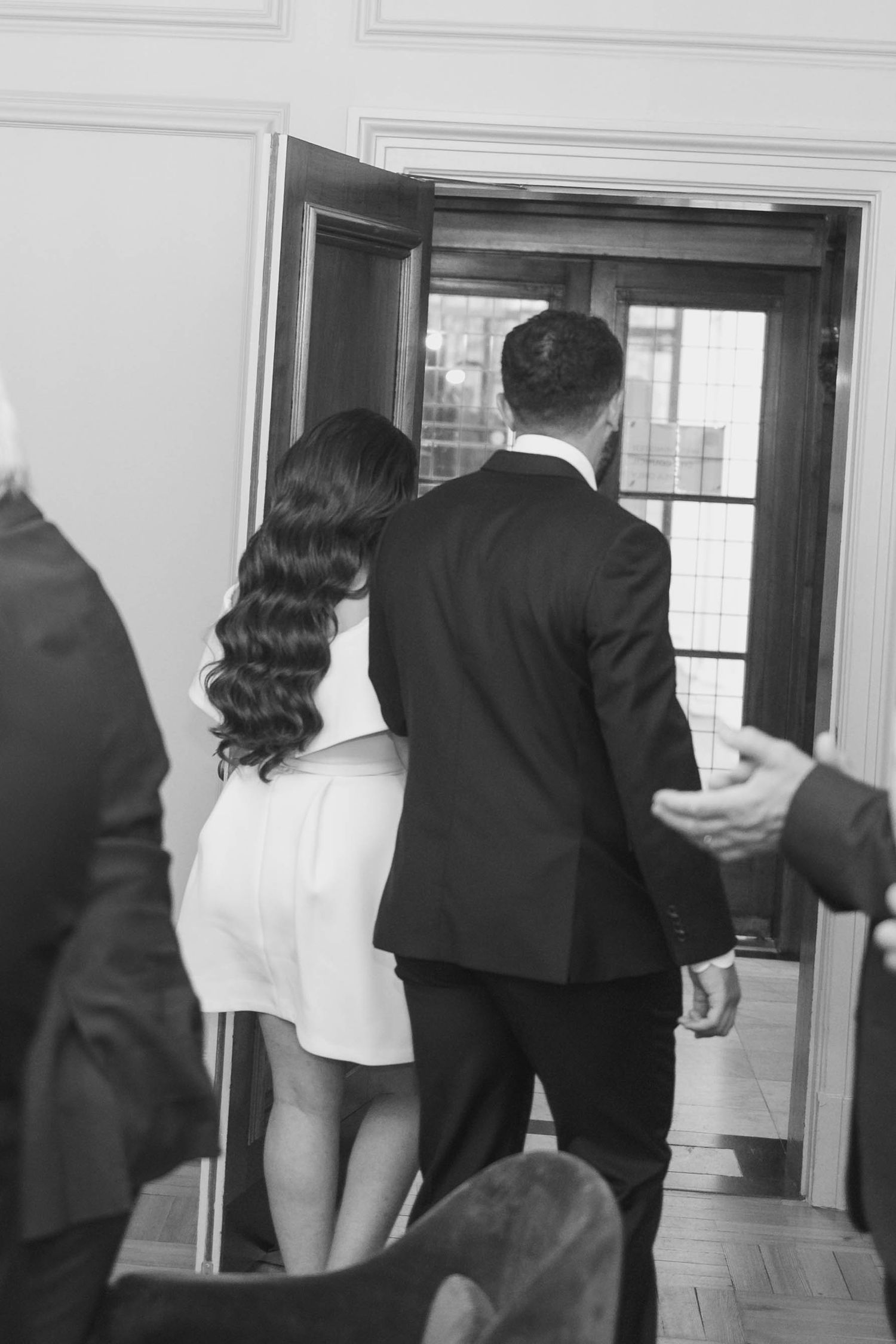 Black and white photo of a couple in formal attire walking through a doorway together.