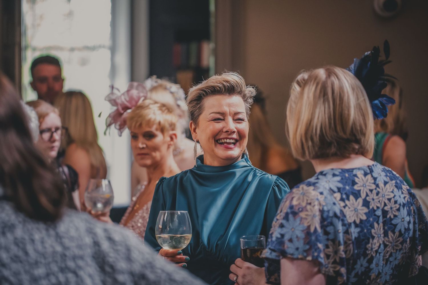 A laughing woman in teal dress enjoys conversation with guests at a wedding cocktail hour.