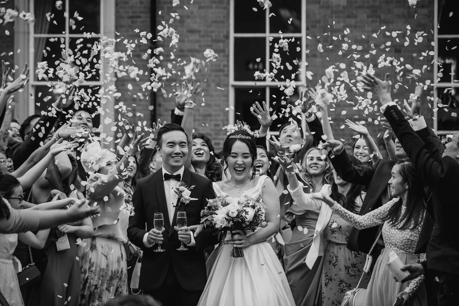 Newlyweds celebrate with guests as white confetti rains down outside their wedding venue in black and white.