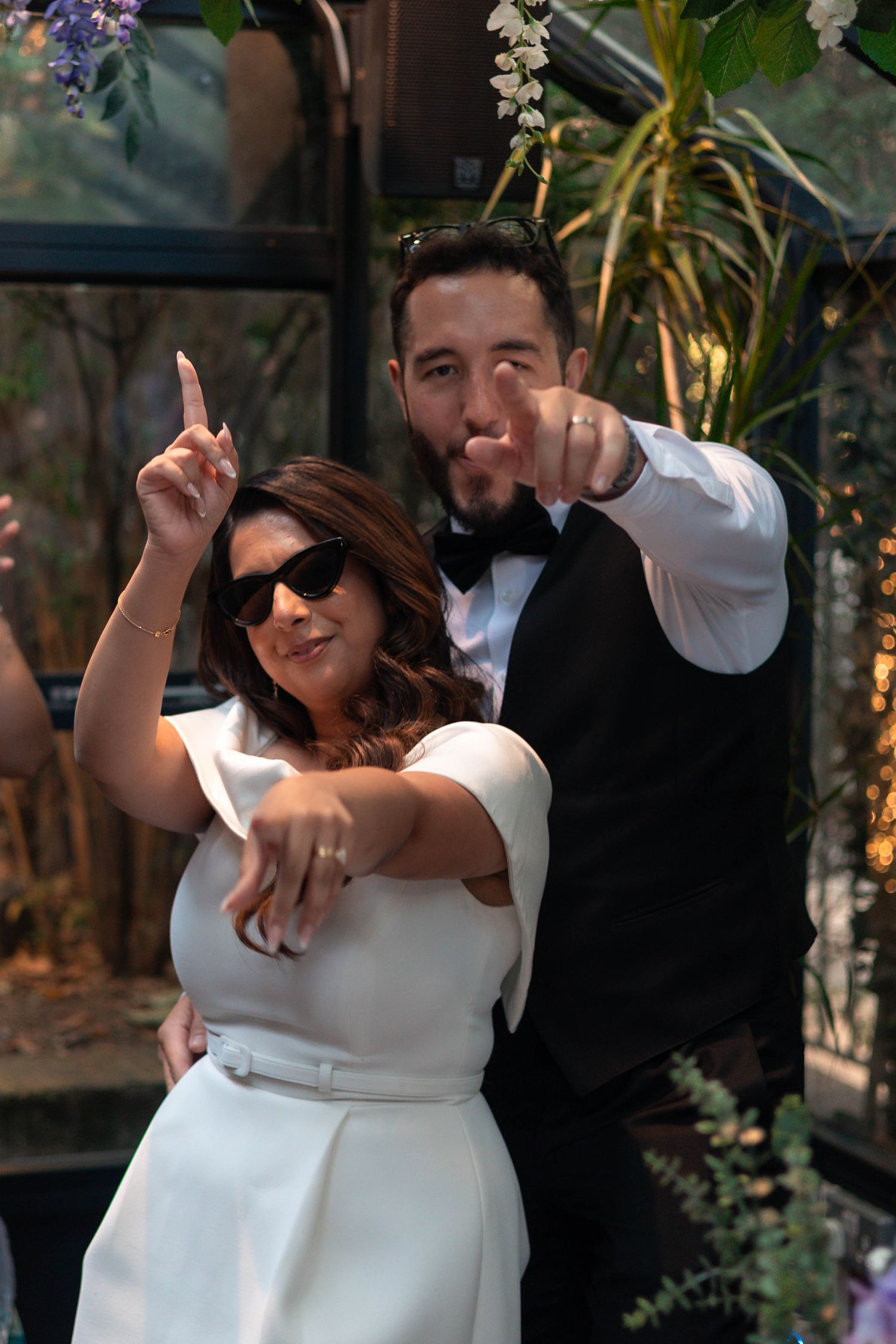 Bride in white dress and sunglasses poses with groom in tuxedo, both pointing at camera at wedding celebration.