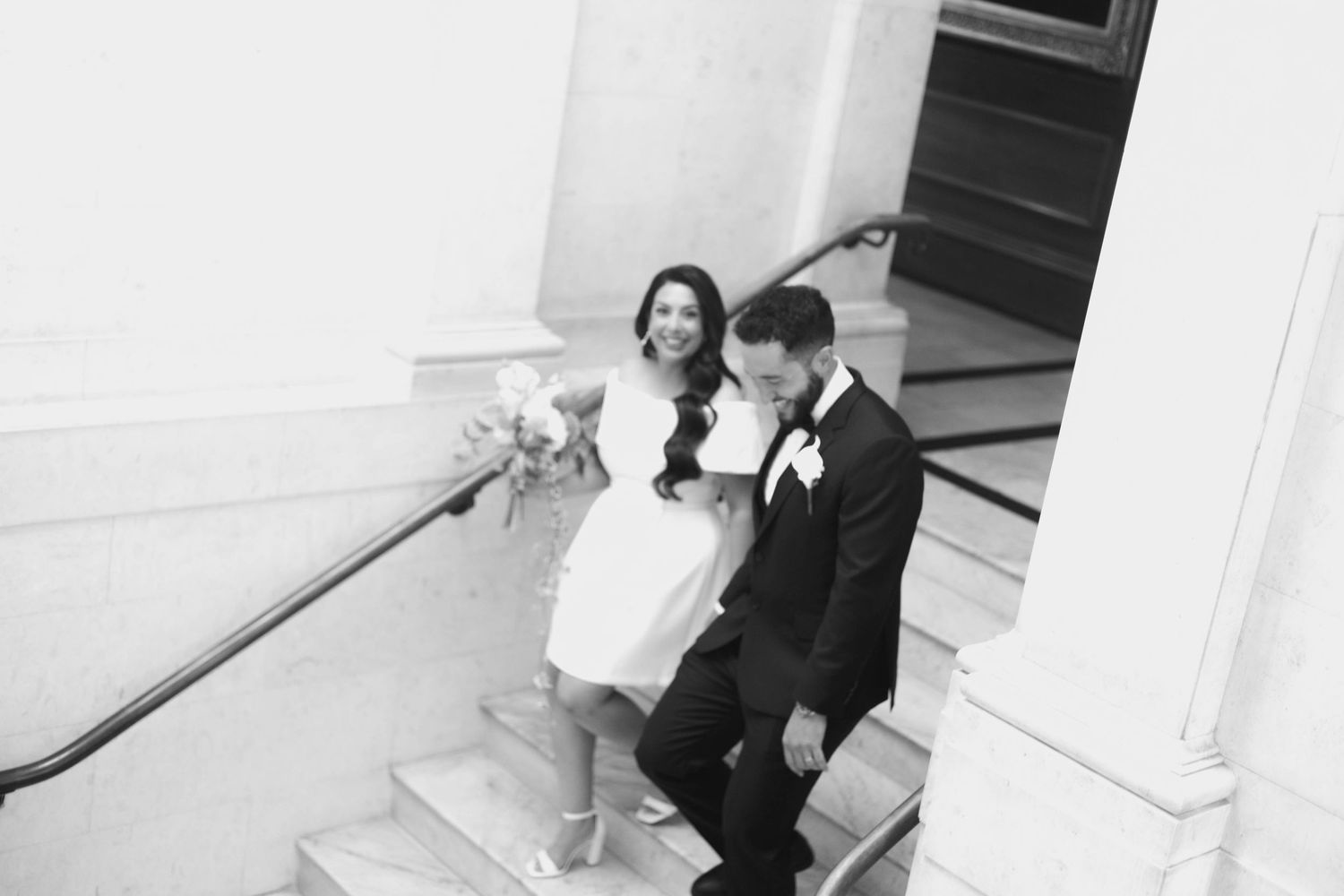 Black and white photo of a bride and groom laughing together while descending elegant stone stairs on their wedding day.