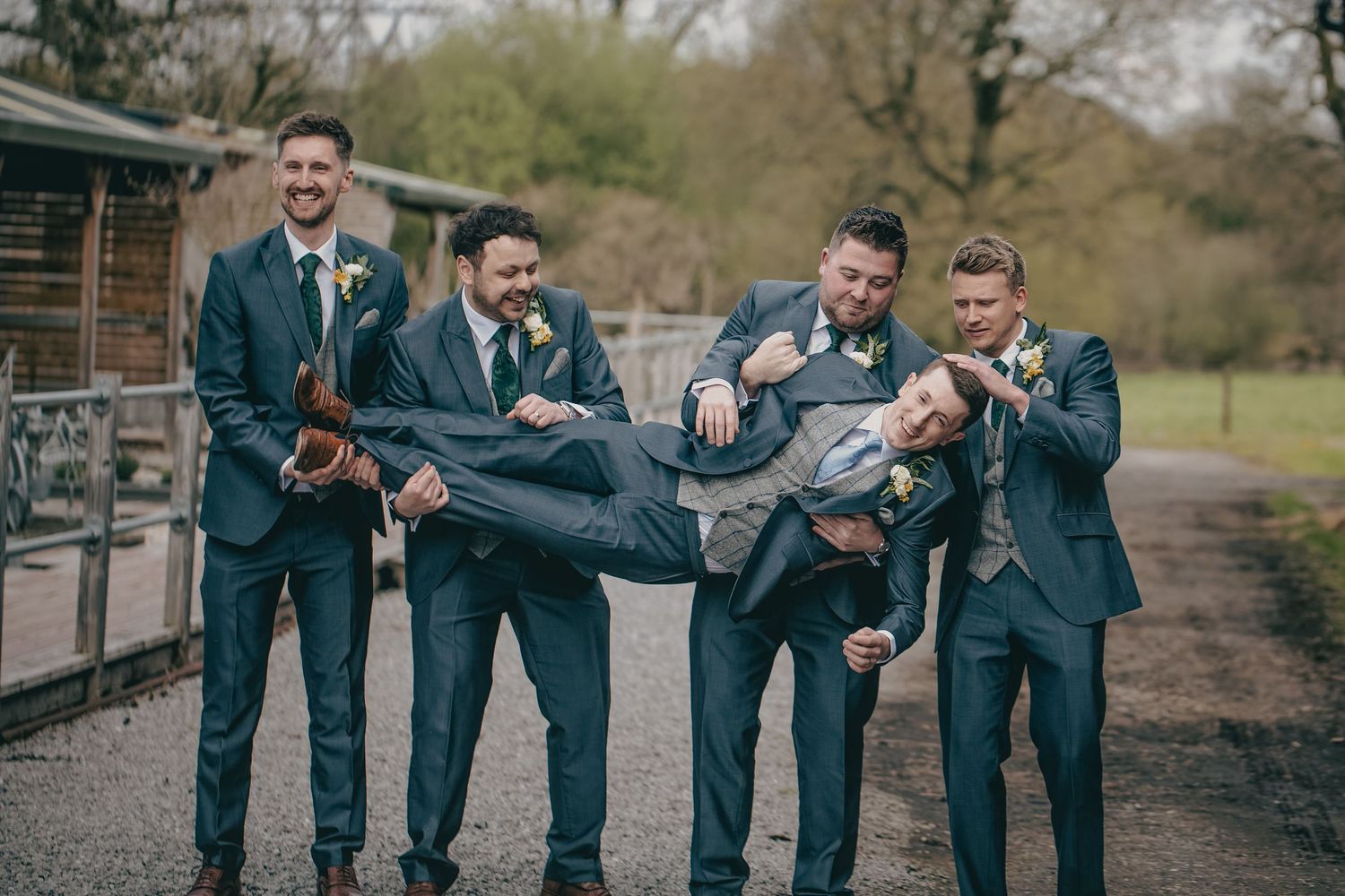 Groomsmen in navy suits laughing while lifting groom horizontally at outdoor wedding venue.