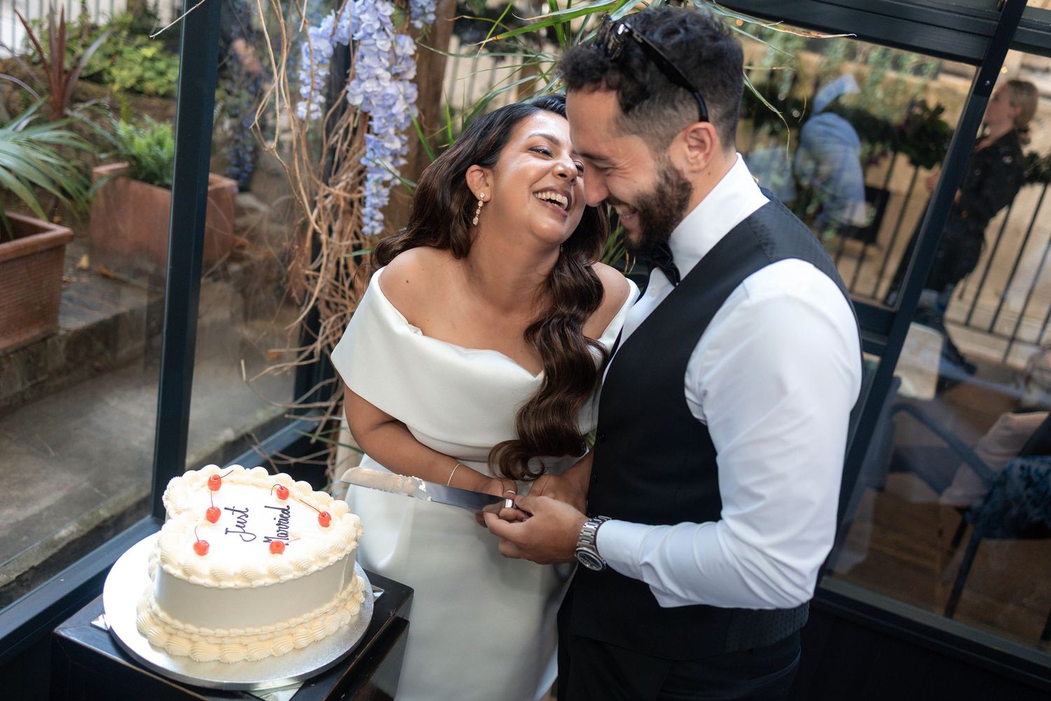 Happy couple laughing together while cutting their white wedding cake at an elegant indoor venue.