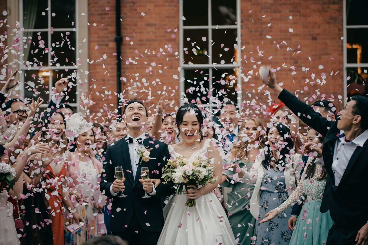 Bride and groom walk through confetti shower with champagne glasses at outdoor wedding celebration.