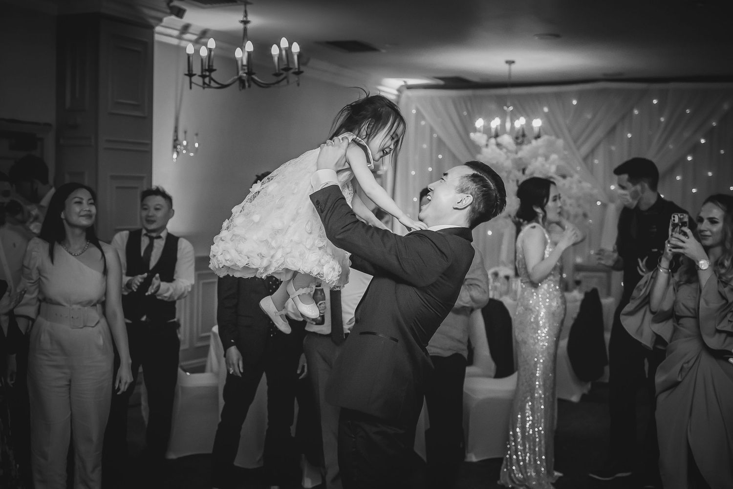 Black and white photo of man lifting young girl in ruffled dress at lively wedding reception.