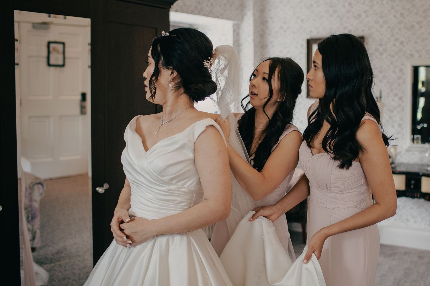 Two bridesmaids in blush pink help a bride in a white satin gown prepare for her wedding.