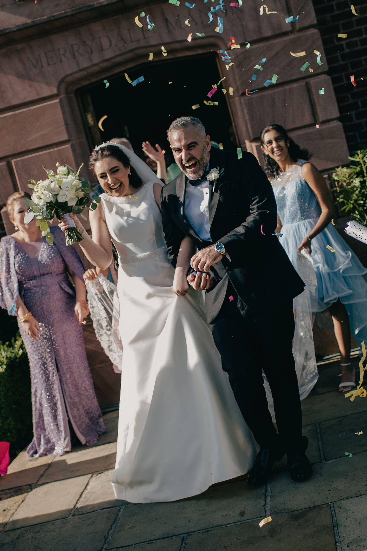 Joyful bride and groom exit church amid colorful confetti shower, surrounded by smiling wedding guests.