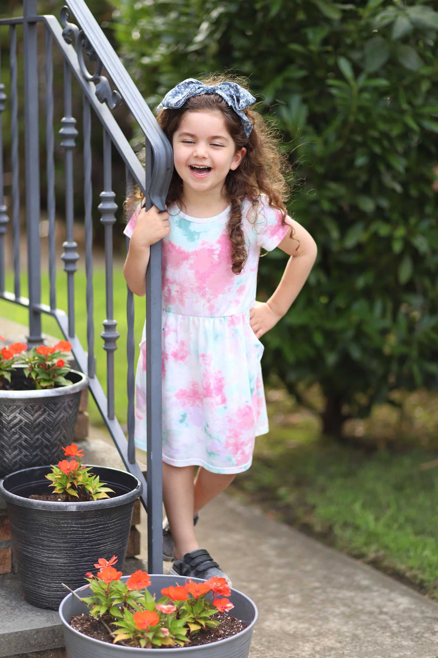 Young girl in tie-dye dress laughing beside stair railing with red flower pots nearby.