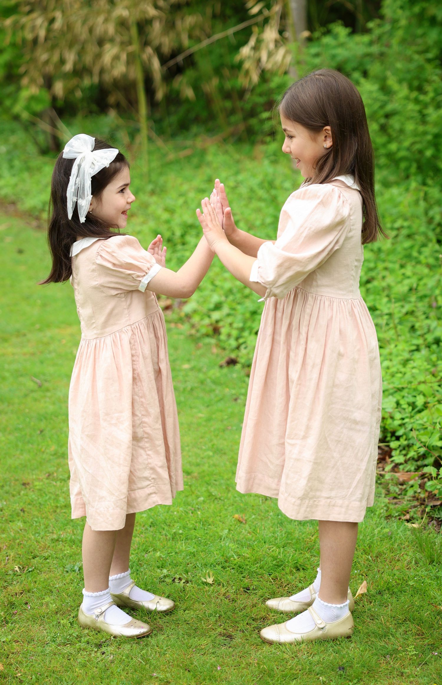 Two young girls in matching pink dresses playing patty-cake outdoors on green grass.
