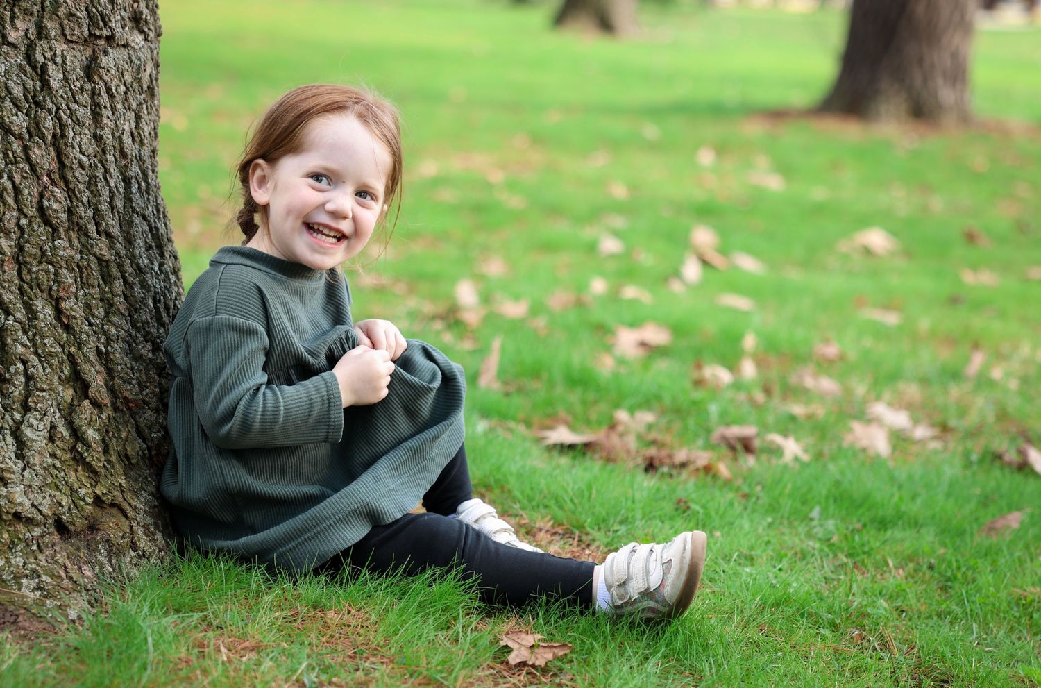 Happy toddler girl sitting against a tree trunk on green grass in a park, smiling and wearing a grey hoodie.