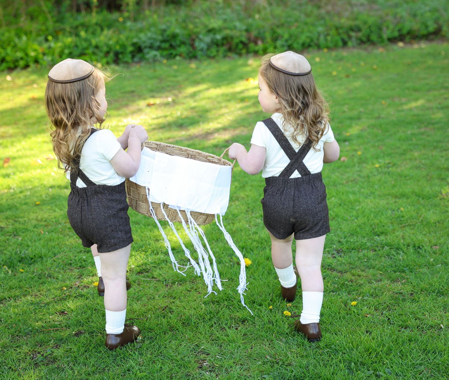Two young children in matching outfits and kippot carrying a basket together on a sunny green lawn.