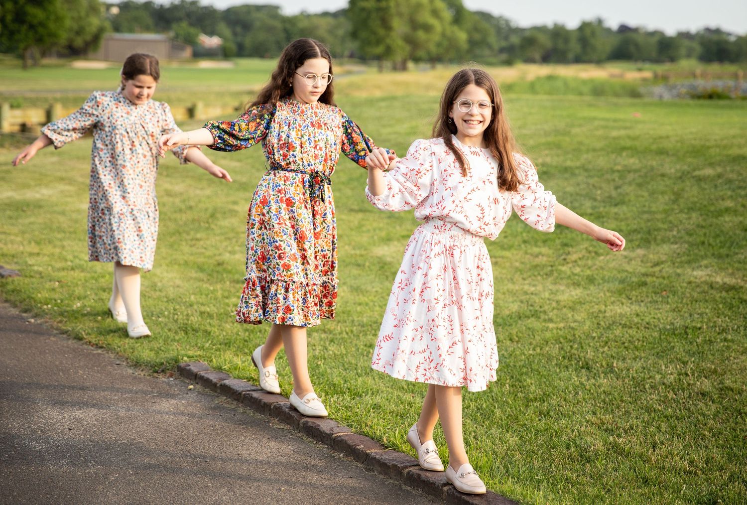 Three girls in floral dresses walk along a path holding hands in a sunny green park.