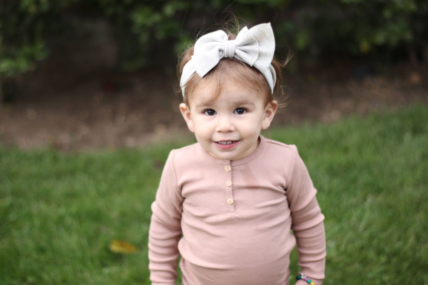 Adorable toddler girl in pink outfit and white bow headband smiling outdoors on green grass.