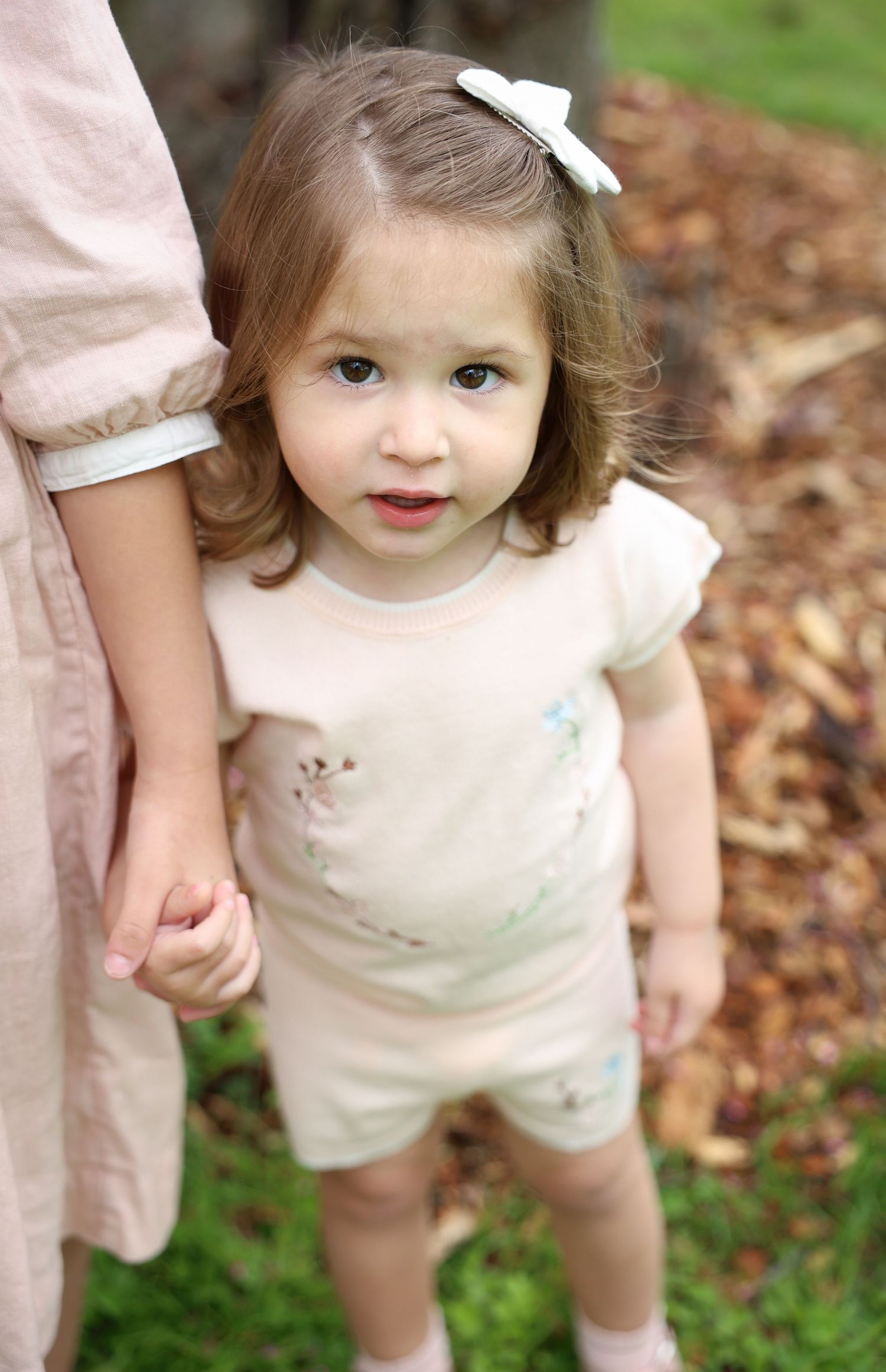 Toddler girl in cream romper with bow hairclip holds adult's hand outdoors among fallen leaves.