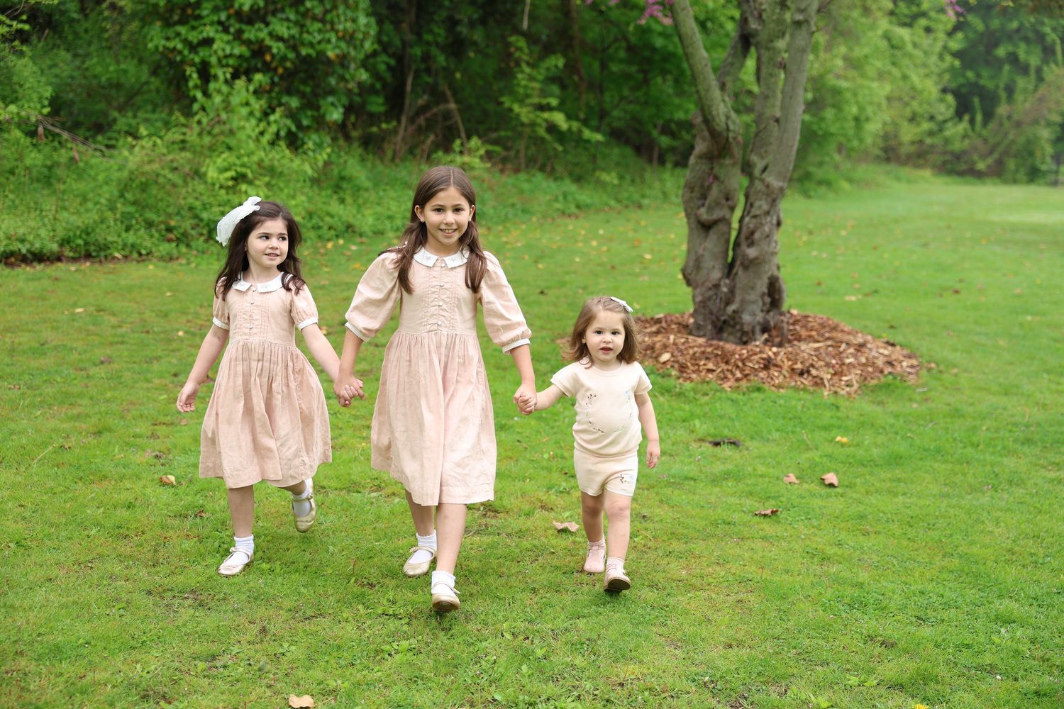 Three young girls in matching pink dresses hold hands while running together on a lush green lawn.