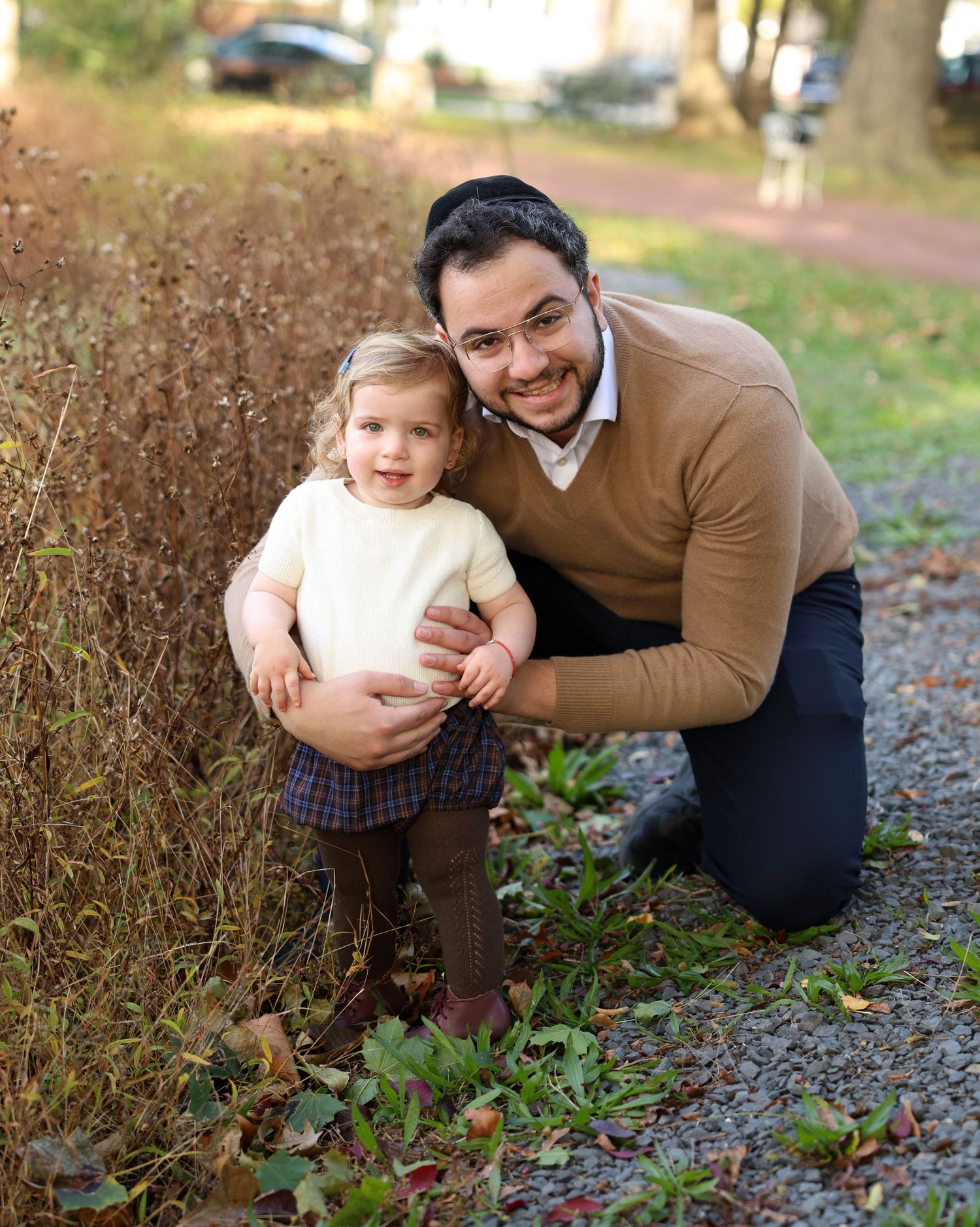 Smiling father kneeling beside toddler daughter in plaid skirt along an autumn garden path.