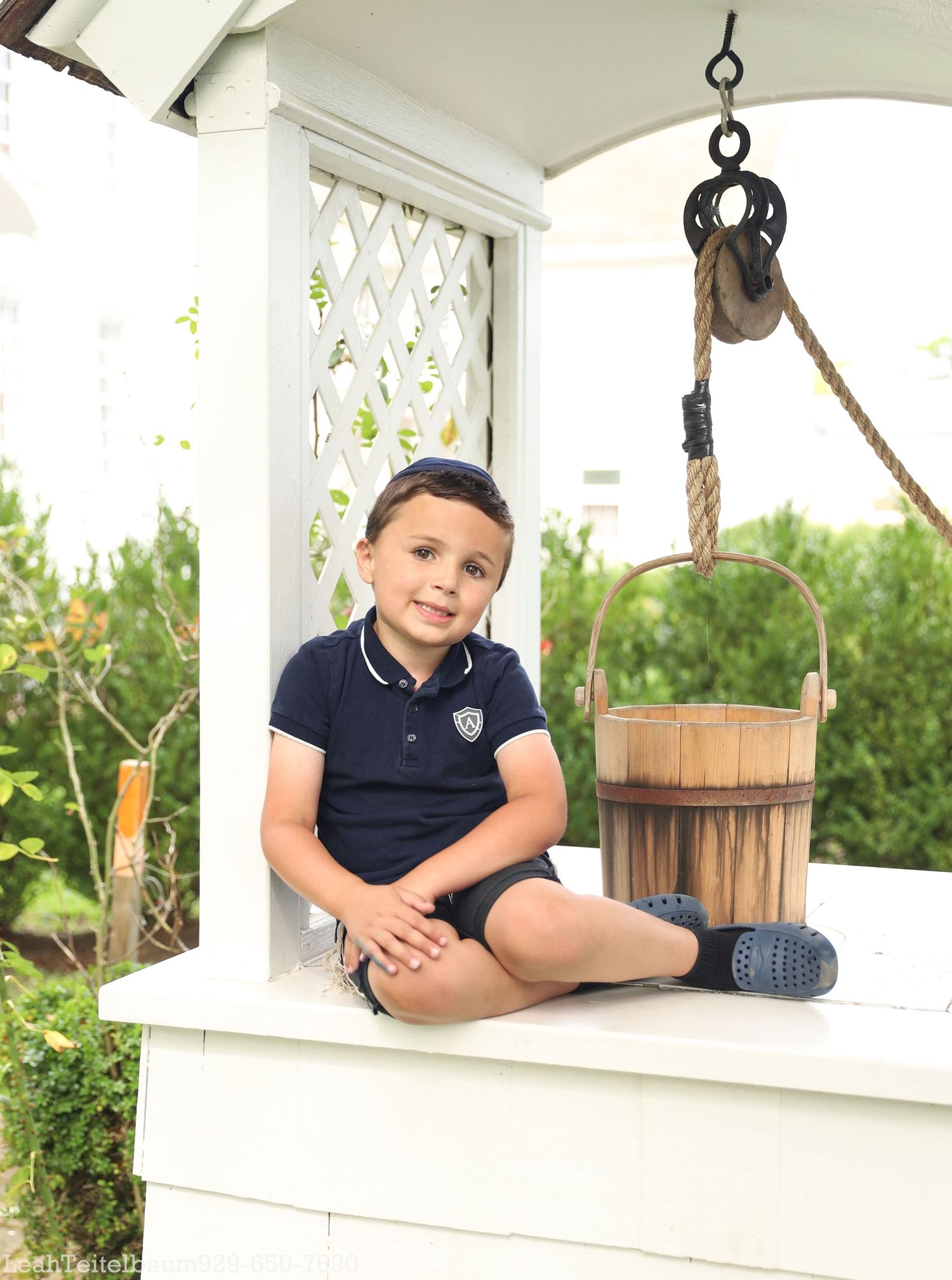 Young boy in navy polo shirt sitting at a decorative white well with a wooden bucket outdoors.
