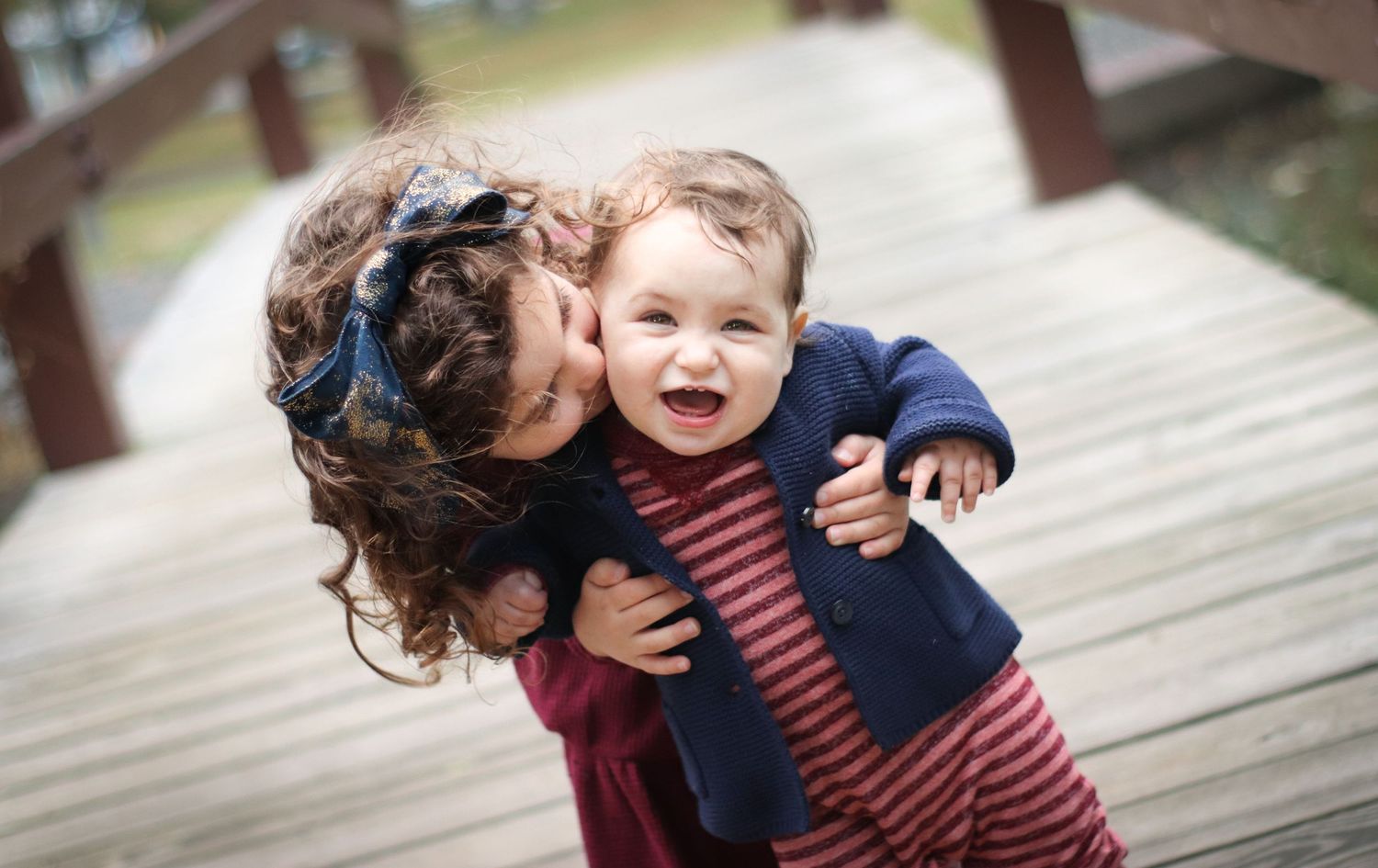Older child hugging and kissing a laughing toddler on a wooden bridge outdoors.