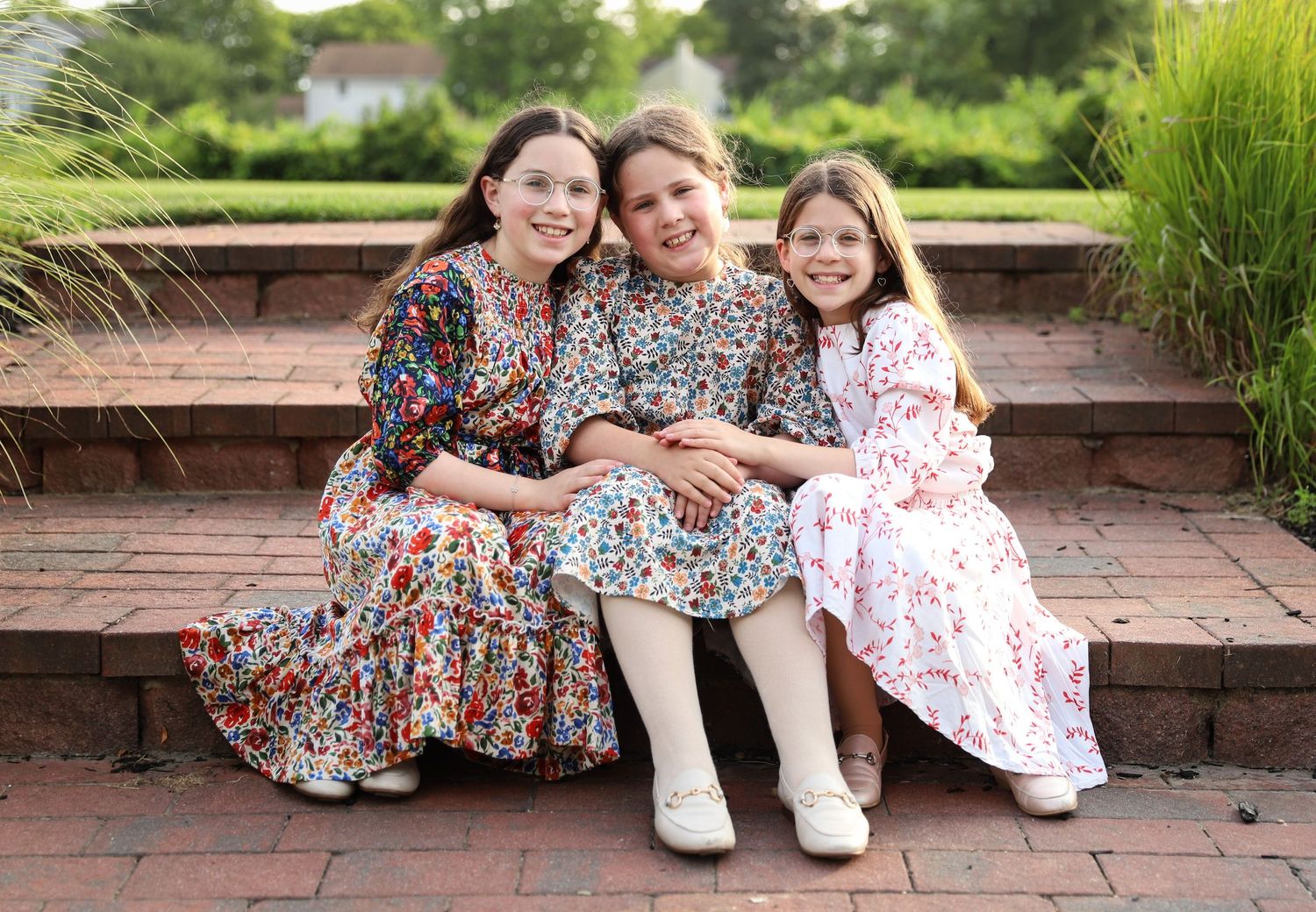 Three sisters in floral dresses sitting together smiling on brick steps outdoors.