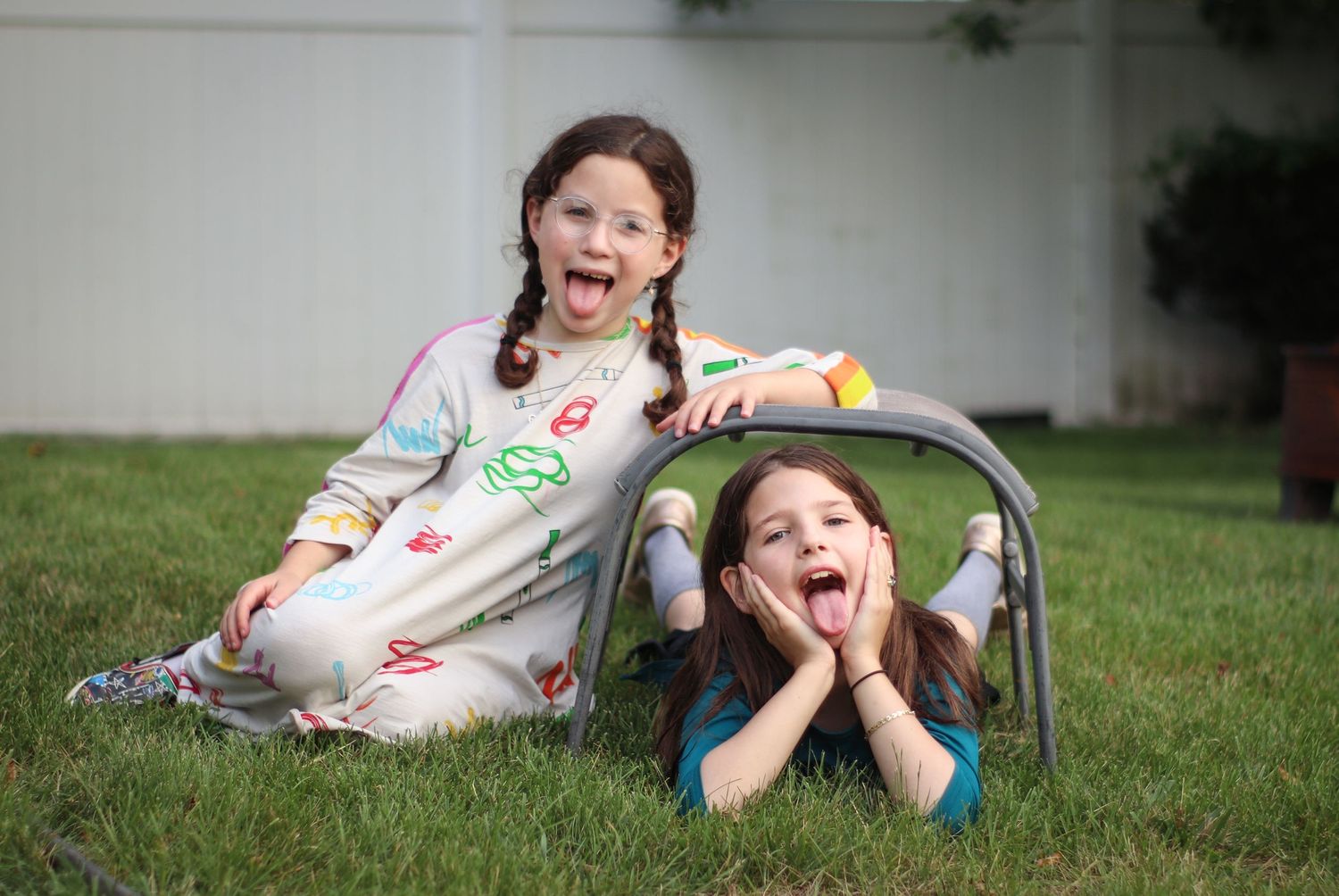 Two happy girls playing outdoors on green grass, one wearing a colorful outfit and the other peeking under a metal arch.