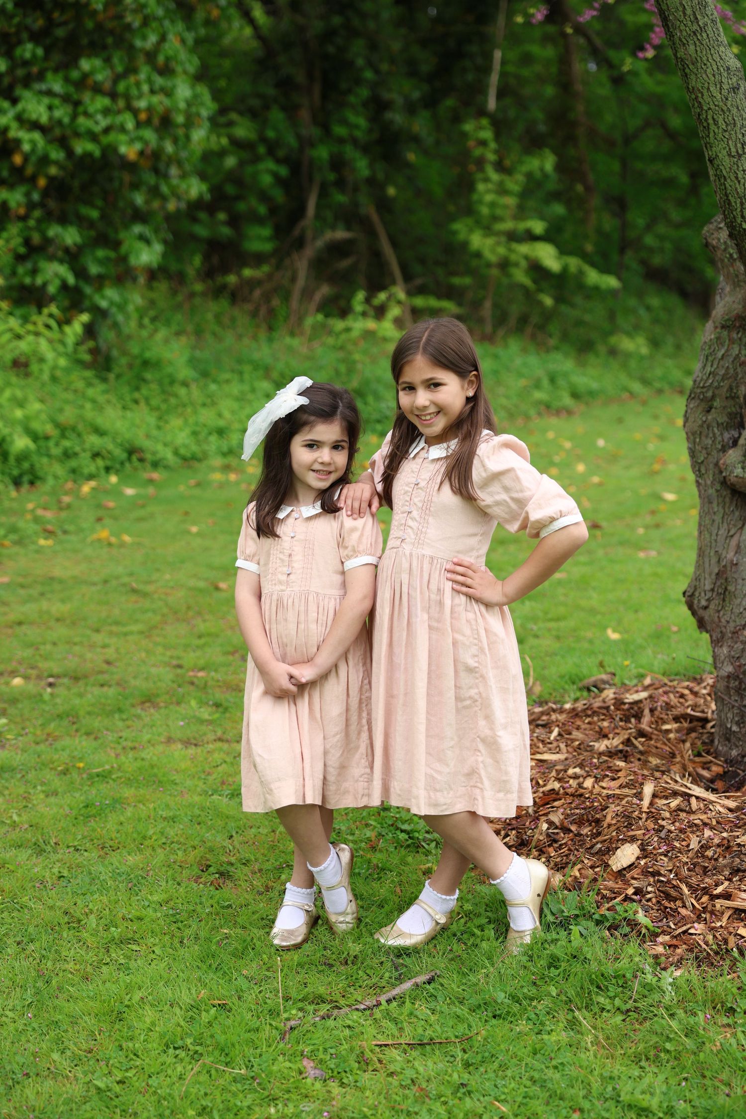Two young girls in matching pink dresses pose together on green grass near a tree in an outdoor garden setting.