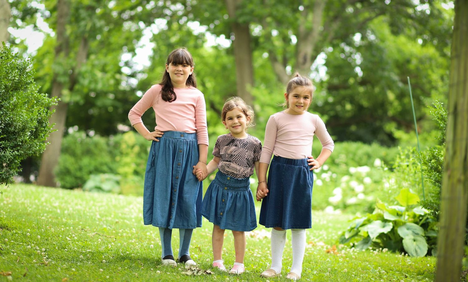 Three young girls in matching pink tops and denim skirts holding hands in a lush green park.