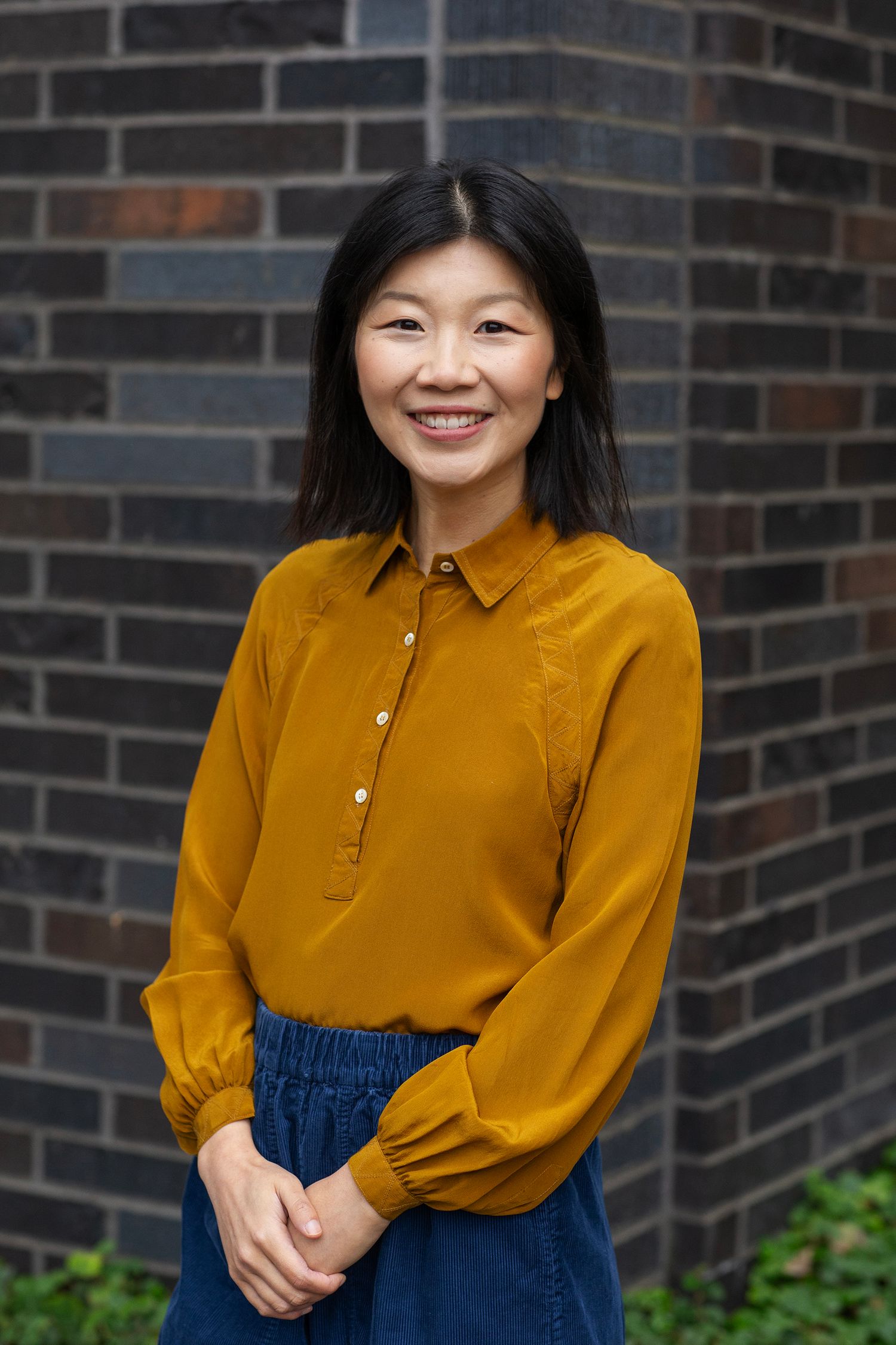 Smiling Asian woman in mustard yellow blouse standing against brick wall outdoors.
