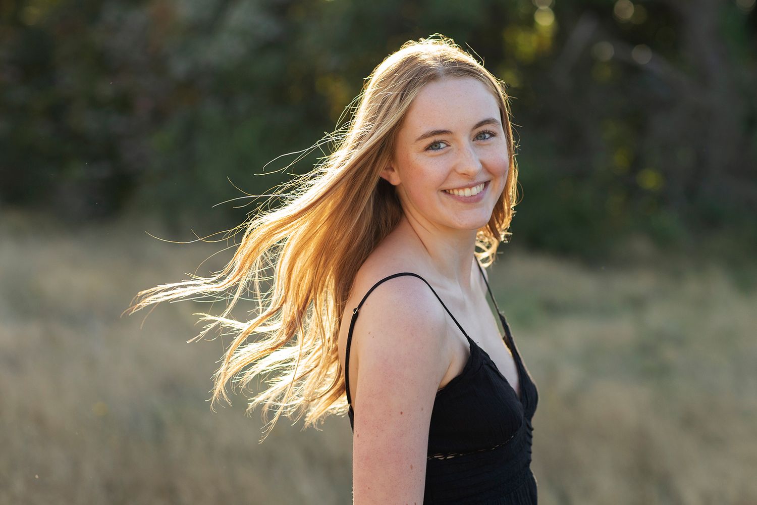 Young woman with long blonde hair smiling outdoors in golden sunlight, wearing a black dress in a natural field setting.