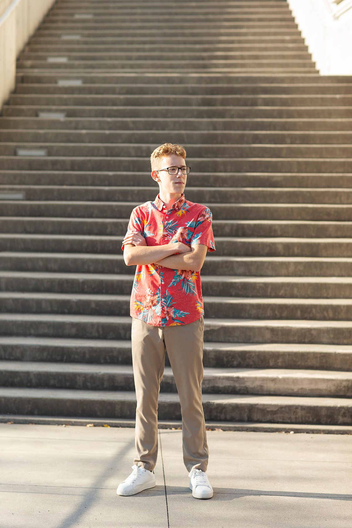 Young man in red floral shirt, khaki pants, and white sneakers stands confidently with arms crossed before stone steps.