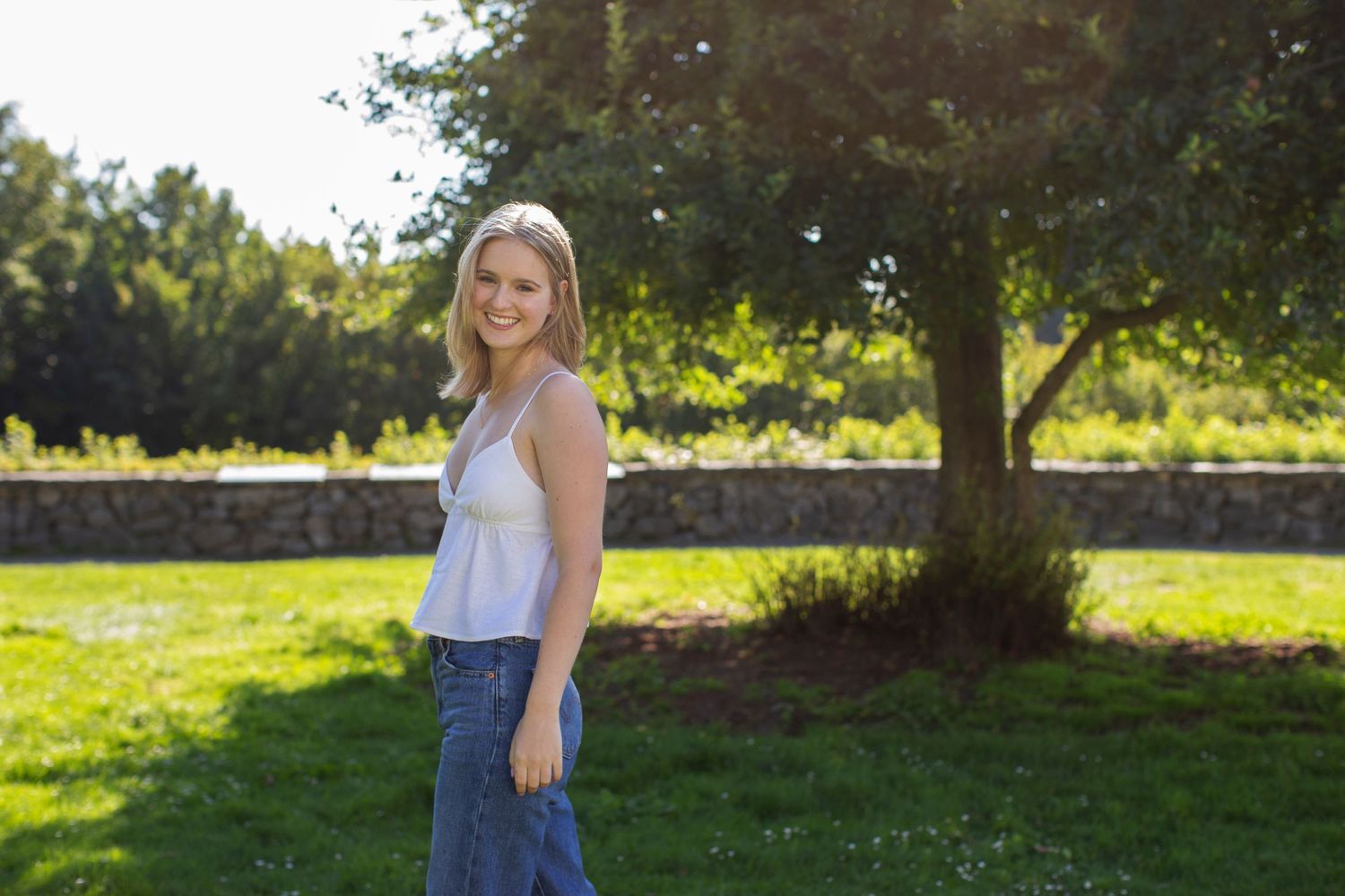 Smiling blonde woman in white cami top and jeans posing in sunny green park with stone wall.