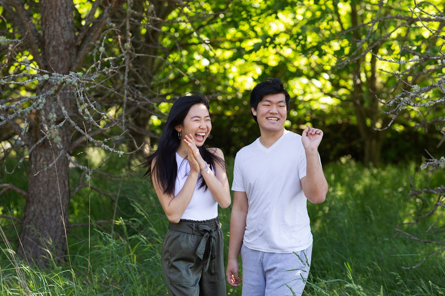 Young Asian couple laughing and walking together outdoors among lush green trees on a sunny day.