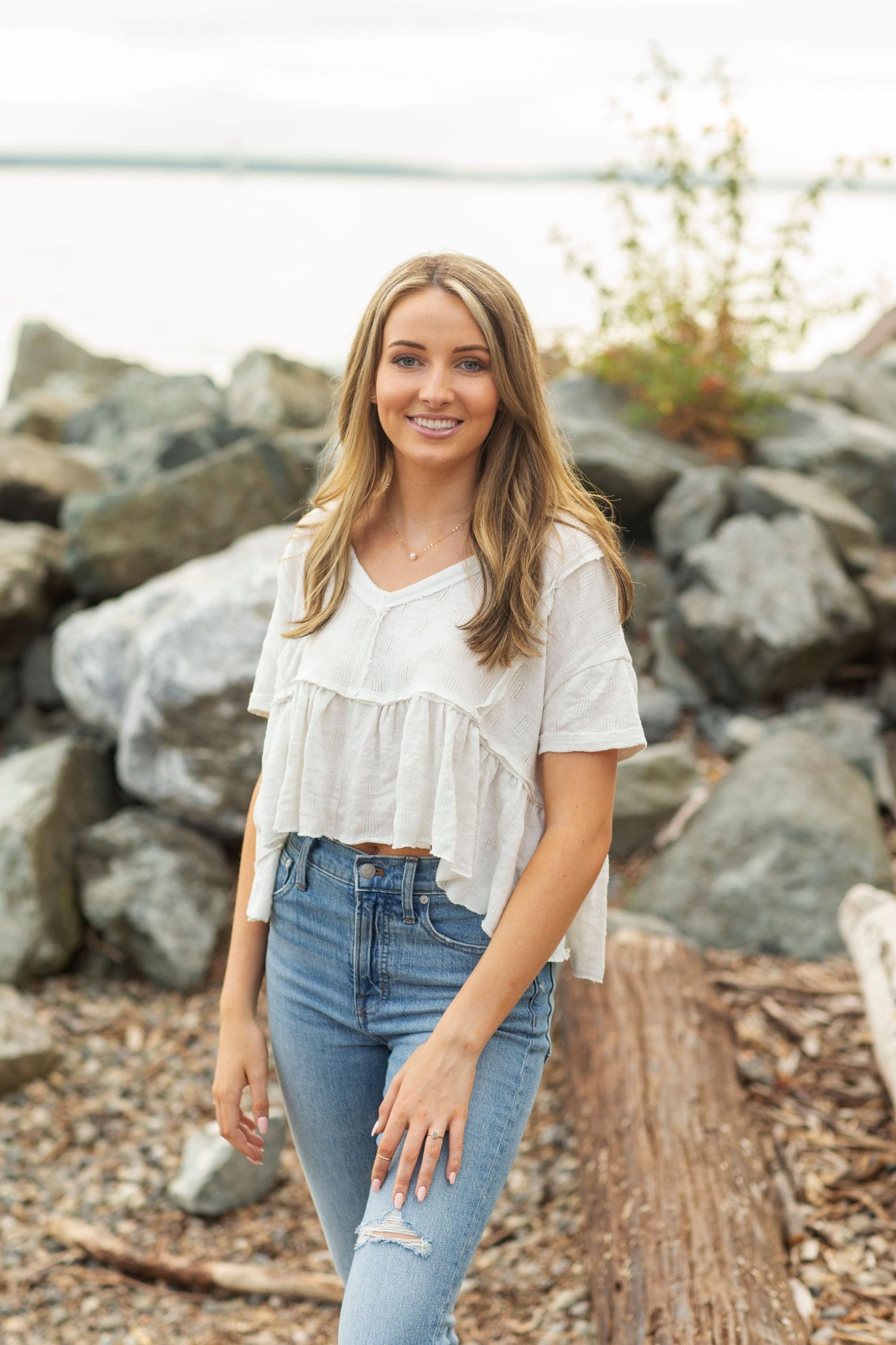 Young woman in white crop top and jeans smiling outdoors near rocky shoreline.