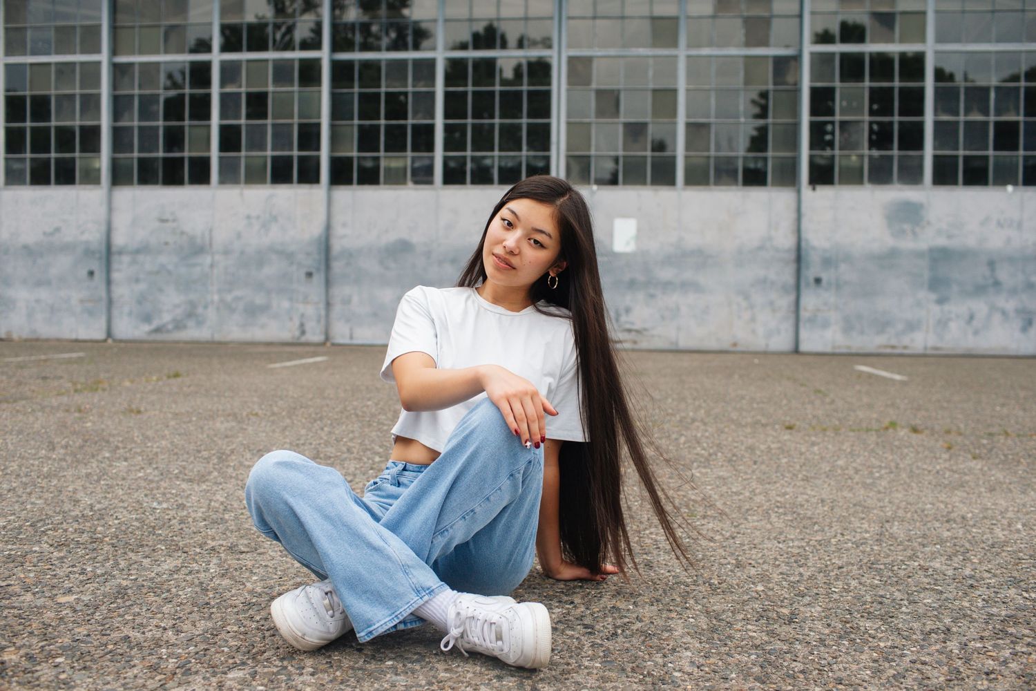 Young woman with long dark hair sitting cross-legged on pavement in white tee and blue jeans.