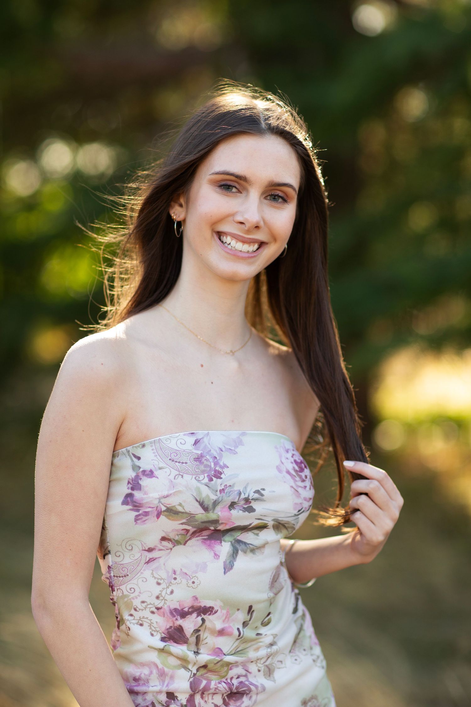 Smiling brunette woman in floral tube dress stands outdoors with sunlit trees in background.