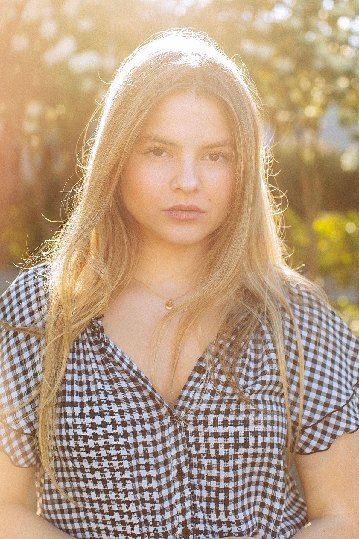 Young blonde woman in checkered shirt poses outdoors in golden sunlight, wearing a delicate moon necklace.