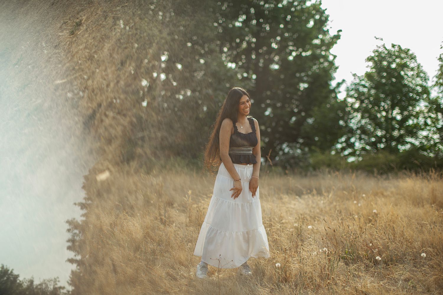 Young woman in black crop top and white skirt standing in sunlit golden field with trees.