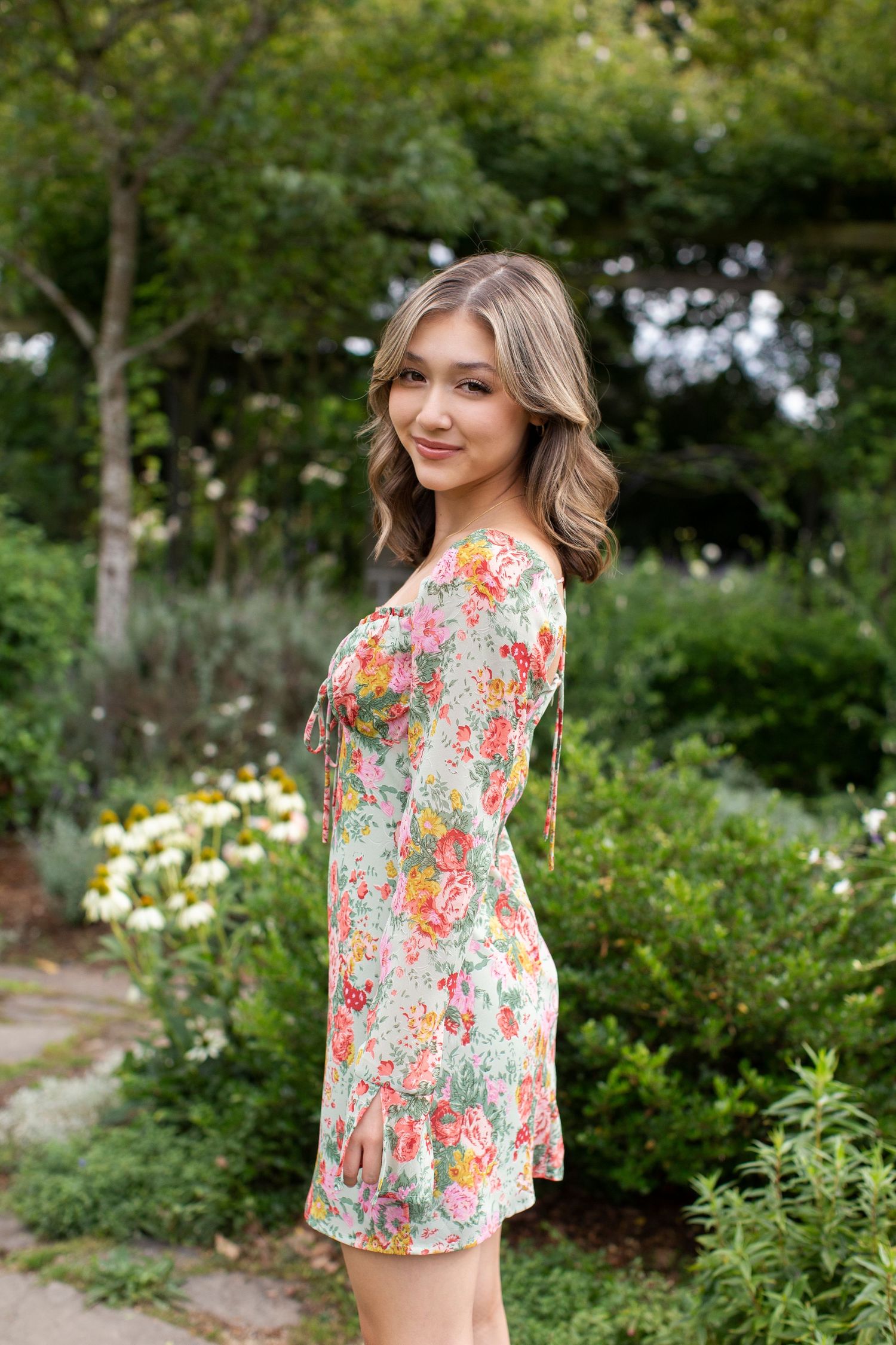 Smiling young woman in a colorful floral mini dress posing sideways in a vibrant green garden.