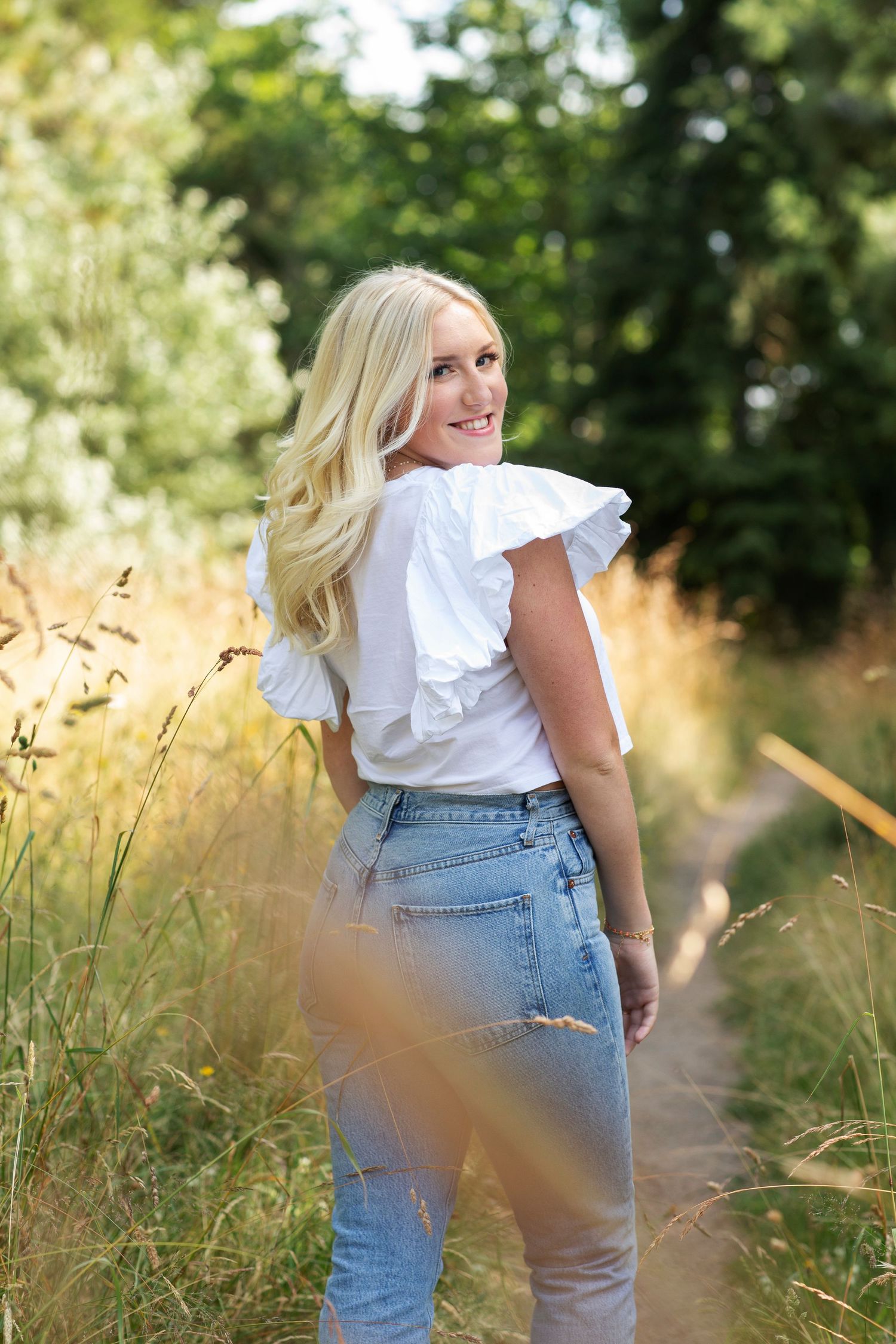 Smiling blonde woman in white ruffled top and jeans on a sunlit nature trail path.