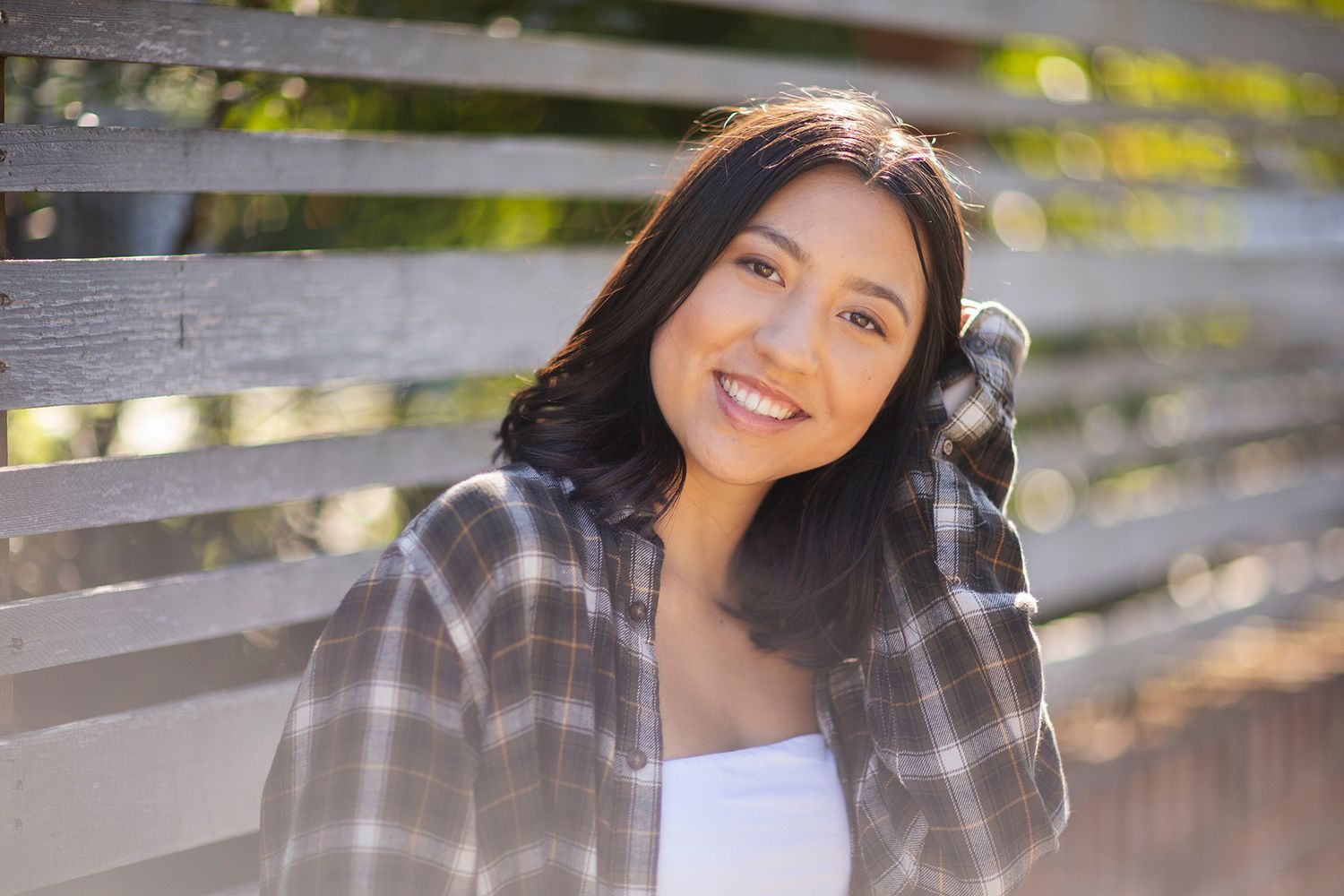 Smiling young woman with dark hair in plaid shirt posing outdoors near wooden fence in natural sunlight.