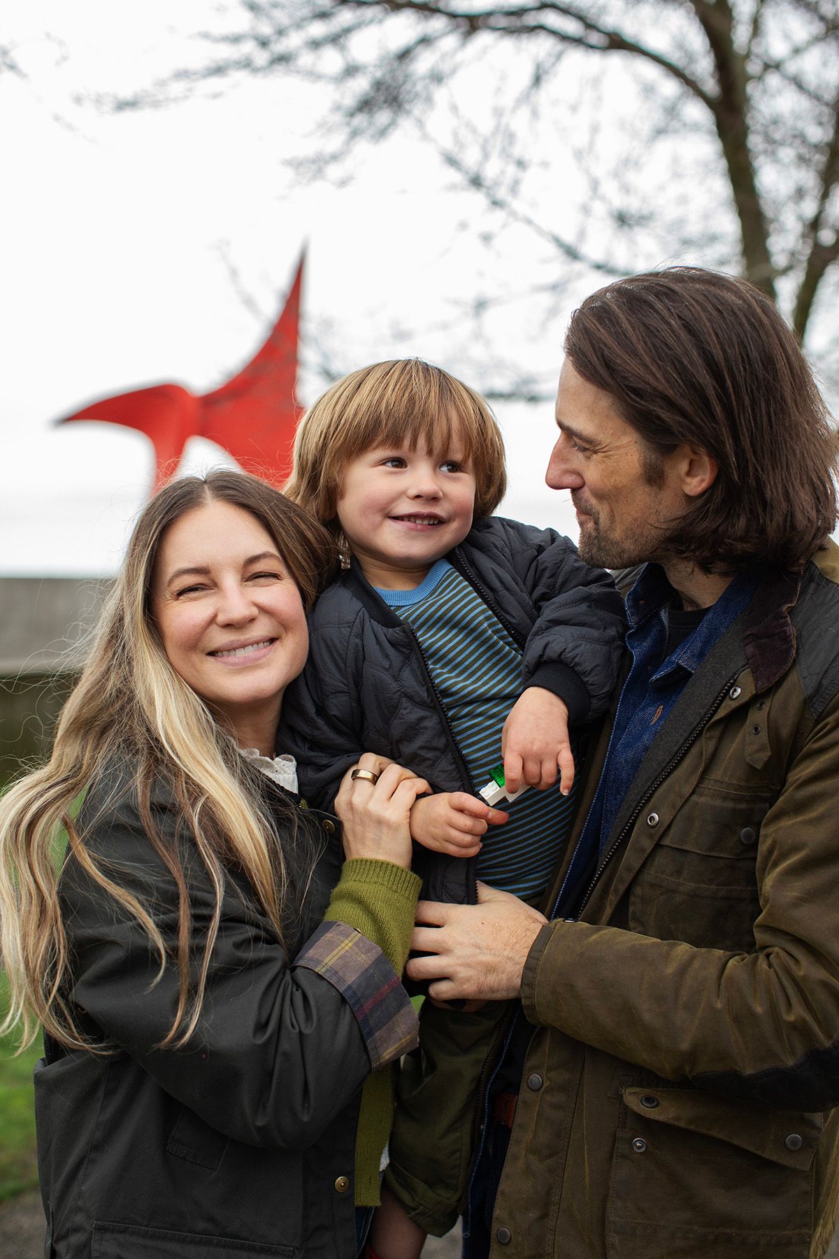A smiling family with a young boy held by his parents outdoors near a red sculpture on a cloudy day.