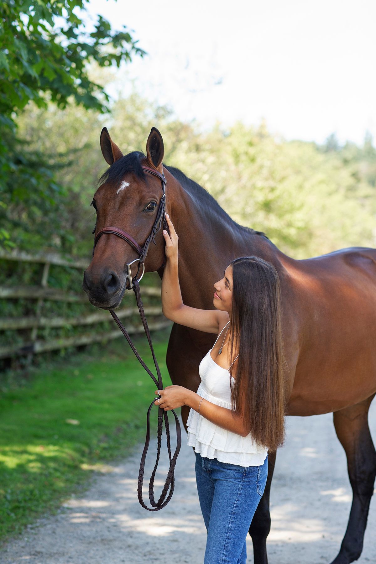 Young woman with long hair gently stroking a brown horse while holding reins on a sunny rural path.
