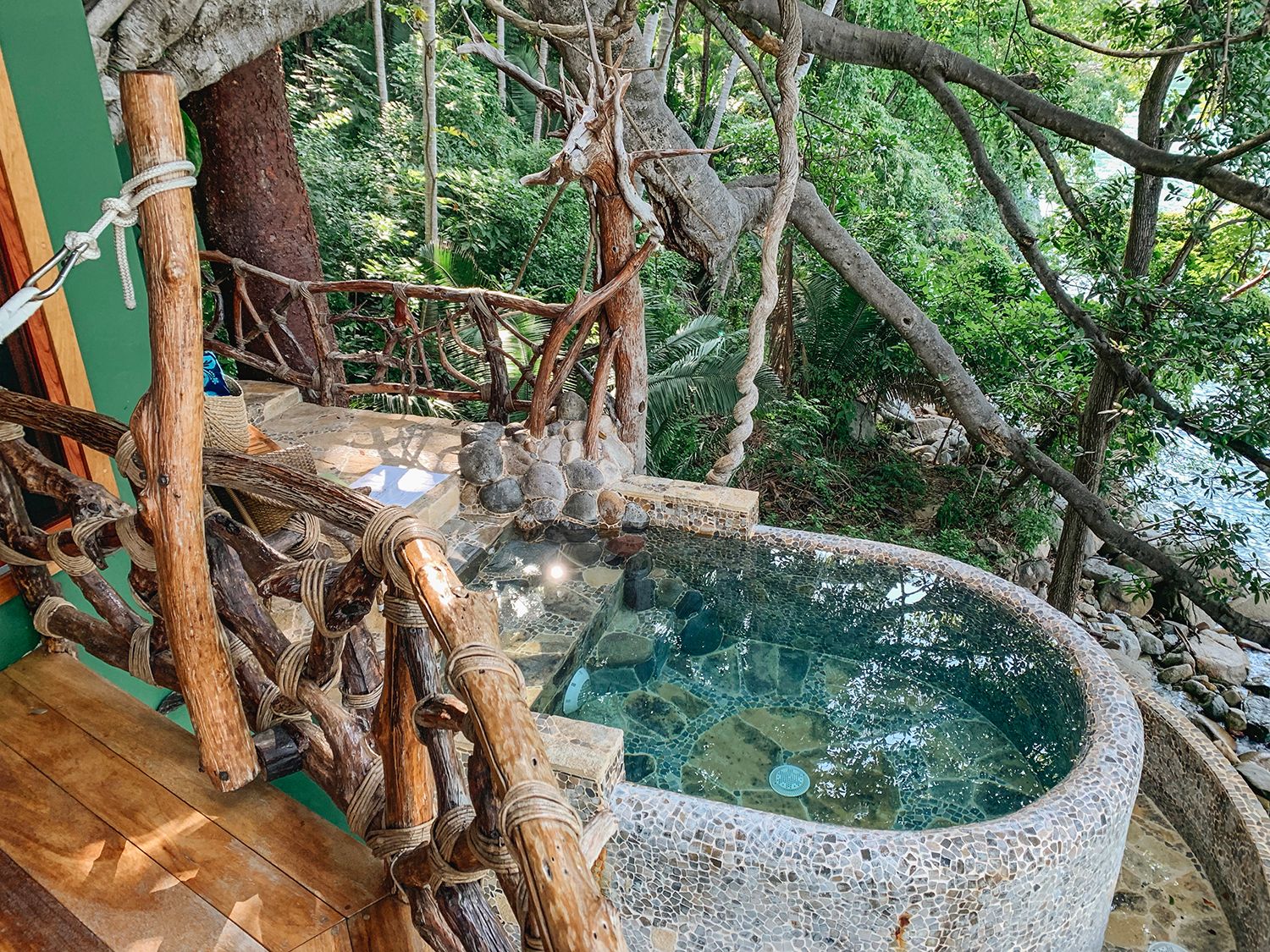 Rustic stone hot tub nestled among twisted tree branches on a jungle deck overlooking lush greenery.
