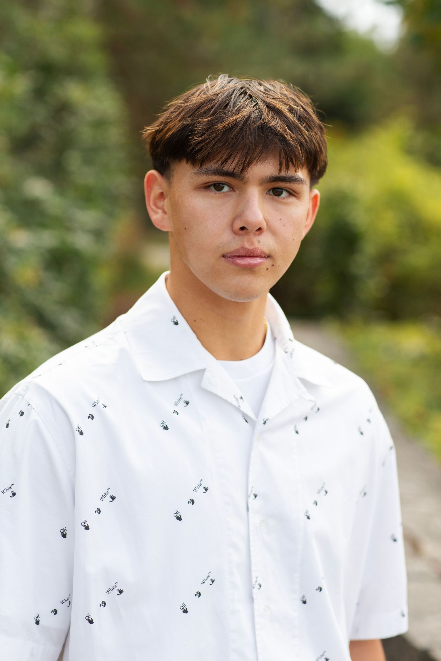 Close-up portrait of teen boy in white short-sleeve patterned shirt outdoors with greenery.