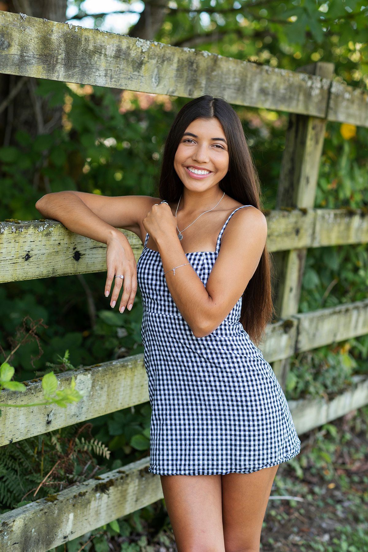 Smiling young woman in gingham dress leaning against rustic wooden fence outdoors.