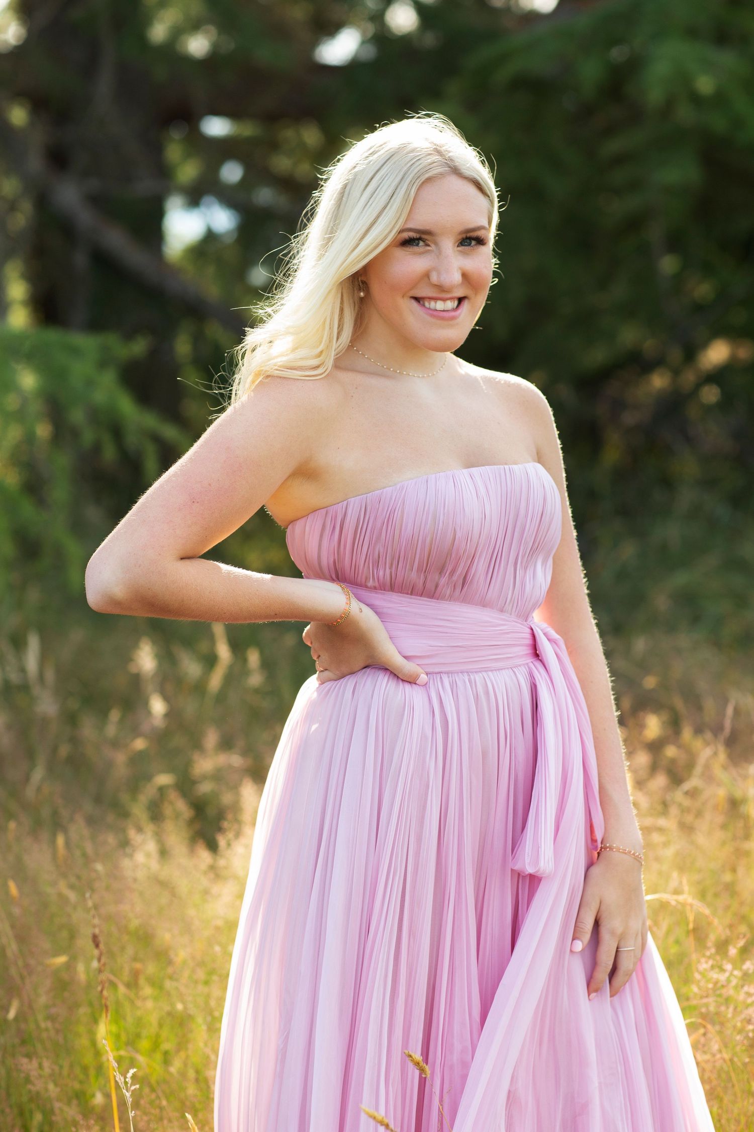 Smiling blonde woman in a strapless pink pleated gown with bow belt posing outdoors in a sunlit meadow.