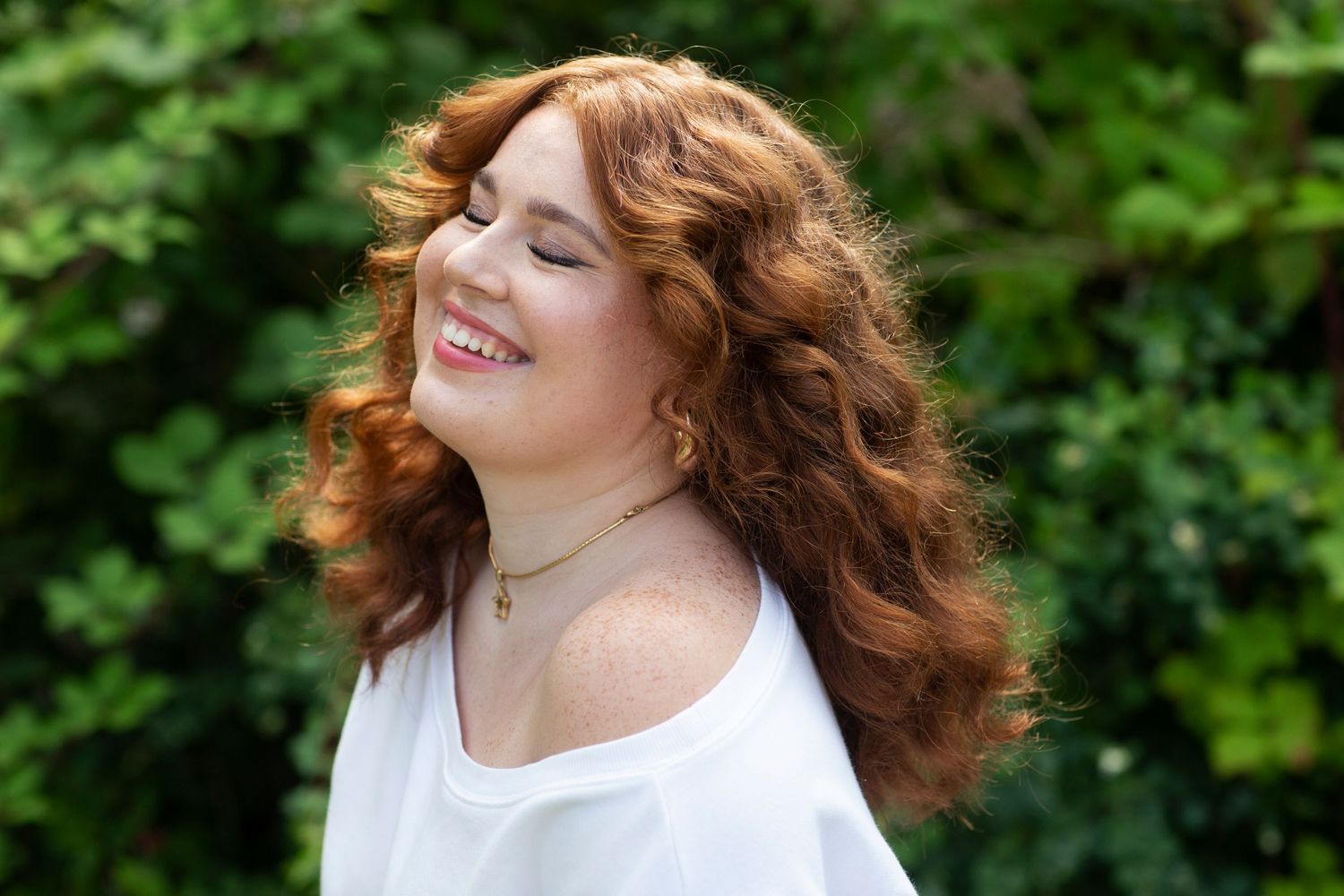 Happy young woman with curly red hair smiling with eyes closed against a green leafy background.