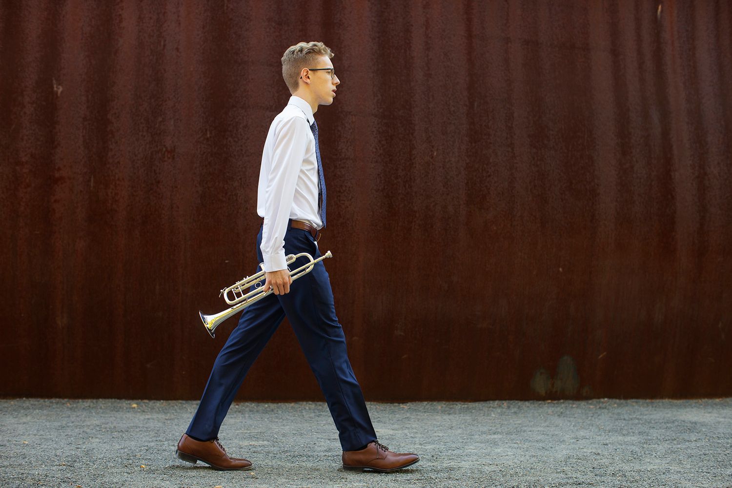 Man in white shirt and blue pants walks while holding a brass trumpet against a rusty red wall.