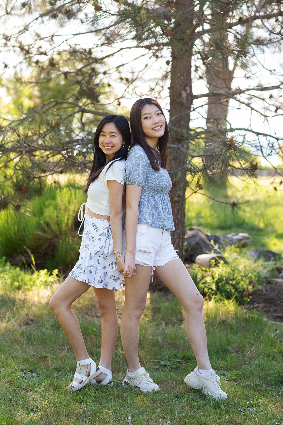 Two young Asian women stand back-to-back holding hands, smiling outdoors near pine trees on a sunny day.